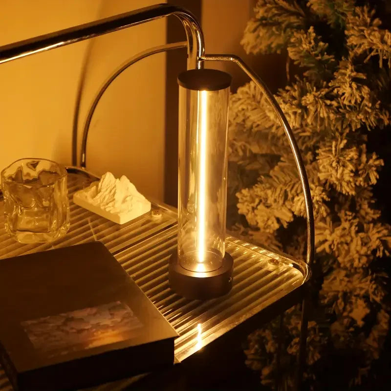 Metallic cylindrical lamp on a metal stand with a warm glow, next to a decorative plant.